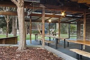 An outdoor covered picnic area with benches and tables, surrounded by trees and a wooden building.