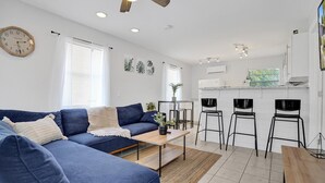 A modern living room with a blue sofa, a wooden coffee table, and a kitchen area with a bar counter and stools.