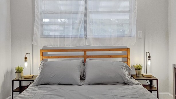 A bed with a wooden headboard, two bedside tables with lamps, and a window with sheer curtains.