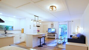 Eine moderne Küche mit weißen Schränken, einer zentralen Kochinsel mit Barhocker, einem Essbereich mit Tisch und Stühlen sowie einem Wohnzimmer mit Sofa und Fernseher.