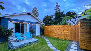 Ein Haus mit überdachter Terrasse, zwei blauen Stühlen, einem Holzzaun und einem Gartenweg.