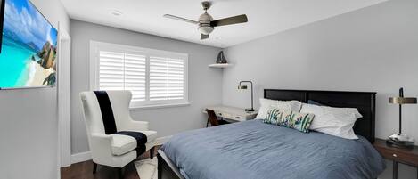 Un dormitorio con cama, escritorio, silla, ventilador de techo y ventana con persianas.