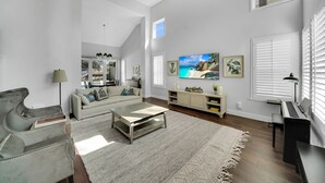 Un amplio salón con sofá, mesa de centro y un piano. Hay un televisor montado en la pared y una ventana con persianas.