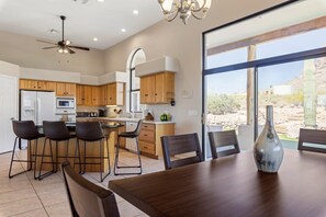 A modern kitchen with wooden cabinets, a central island with bar stools, a dining table with chairs, and a large window offering a view of the desert. - Stunning Views Superstitions Hot Tub Putting Green (Apache Junction)