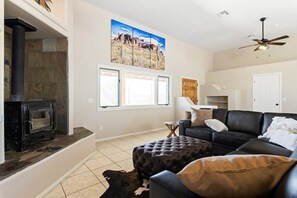 A living room with a fireplace, a black leather sofa, a large painting, and a ceiling fan. - Stunning Views Superstitions Hot Tub Putting Green (Apache Junction)