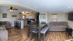 A spacious living area with a large L-shaped couch, a dining table with benches, a kitchen with wooden cabinets, and a ceiling fan.