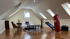 Une chambre spacieuse aménagée en mezzanine, avec un plafond en pente, un plancher en bois, un lit, un bureau, une chaise, un sac de frappe et une fenêtre équipée de stores.