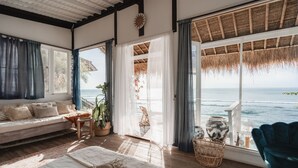 Quarto espaçoso com uma grande janela com vista para o mar, uma área de estar acolhedora com um sofá e cadeiras, e teto de palha.
