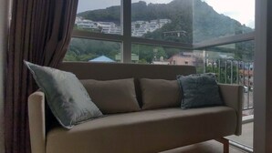 A sofa with cushions in a room with large windows offering a view of a hillside with buildings. - Kata Beach Ozone Superb Fully Equipped Studio Kitchen Washing Machine air con (Karon)