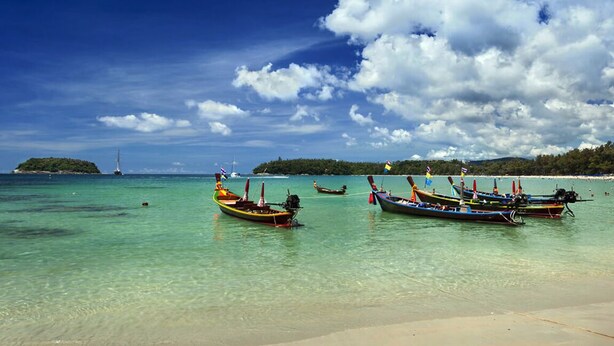 A beach with clear turquoise water, several traditional boats anchored near the shore, and a distant island with a lighthouse. - Kata Beach Ozone Superb Fully Equipped Studio Kitchen Washing Machine air con (Karon)