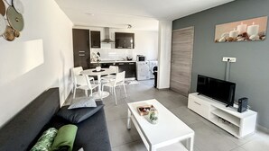 A modern living room with a sofa, coffee table, TV, and a dining area with chairs. - House With Free Parking 15min From The Airport (Charleroi)