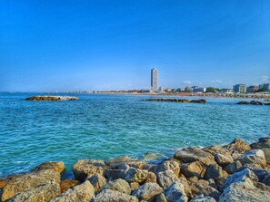 A coastal cityscape with a prominent tall building, rocky shoreline, and a clear blue sky.