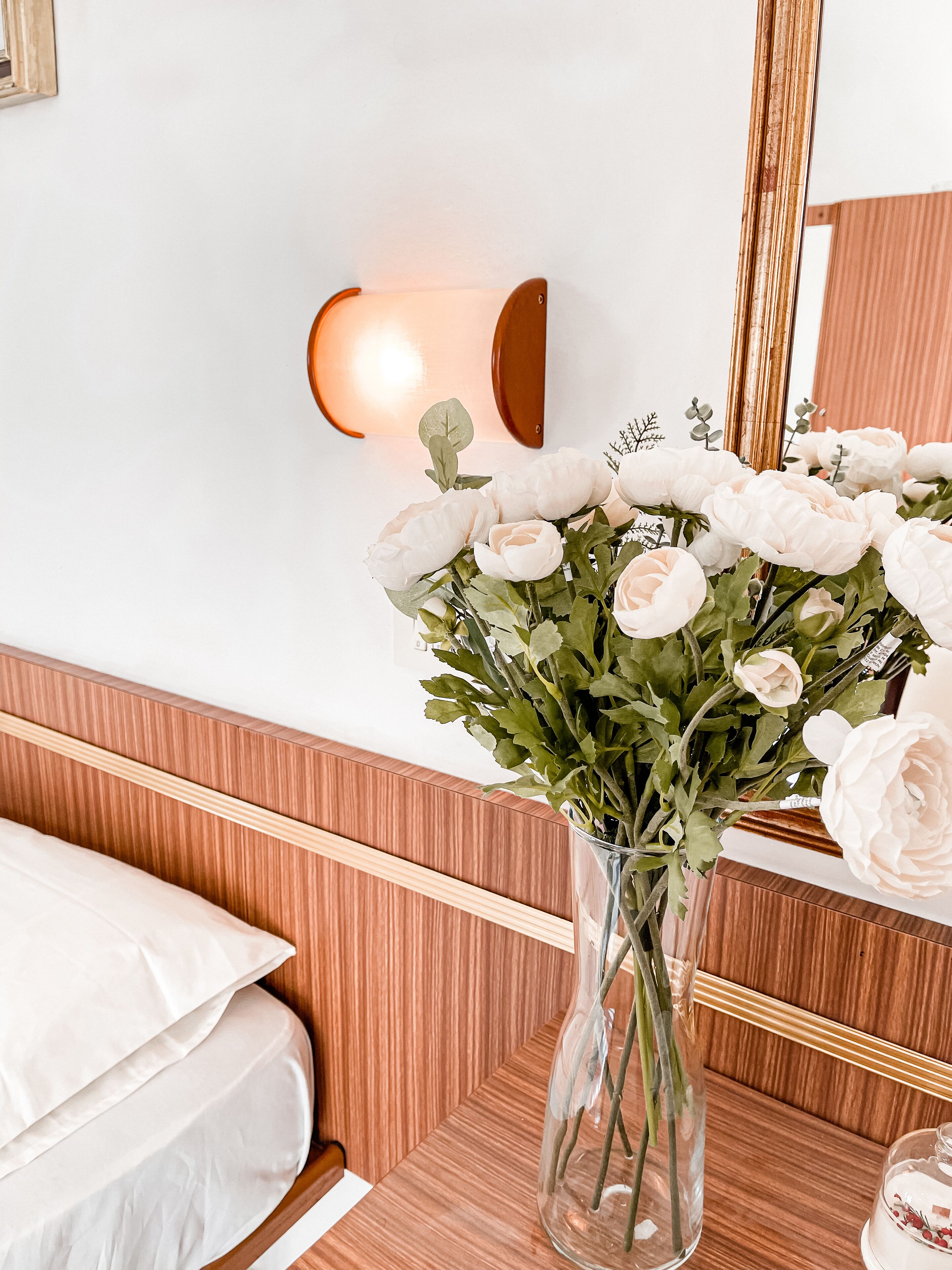 Un dormitorio con cabecera de madera, cama blanca, un florero con flores blancas y una lámpara de pared.