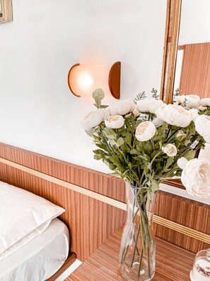 A bedroom with a wooden headboard, a white bed, a vase with white flowers, and a wall-mounted light fixture.