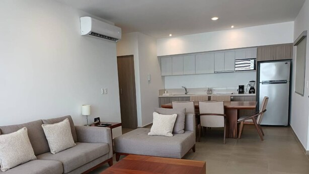 A modern living room with a sofa, ottoman, and coffee table, a kitchen with grey cabinets and a refrigerator, and a dining area with chairs. - One Bedroom, Great Location Steps From the Beachh, Beautiful Roof Top View! (Playa del Carmen)