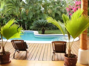Zona de piscina con suelo de madera, dos sillas de madera y plantas en macetas.