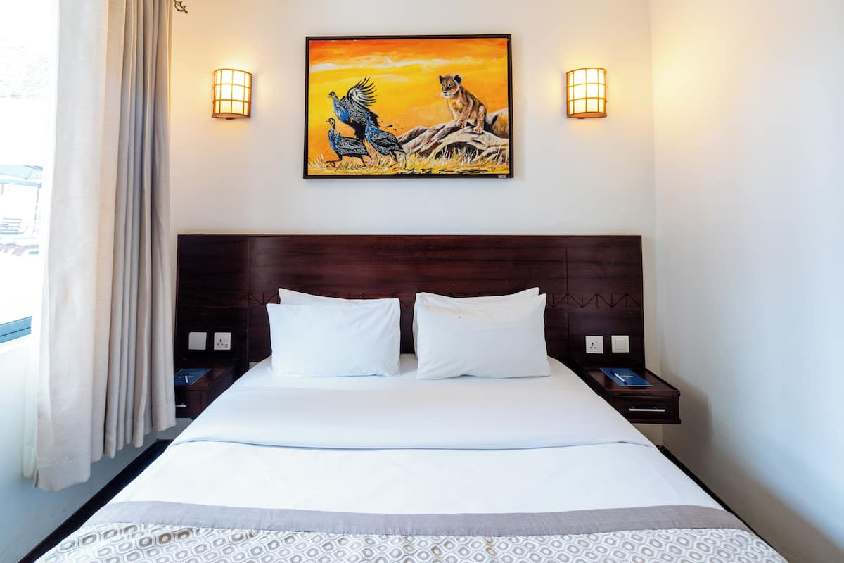 A hotel room with a large bed, two bedside tables, a nightstand, a painting above the bed, and a window with curtains.
