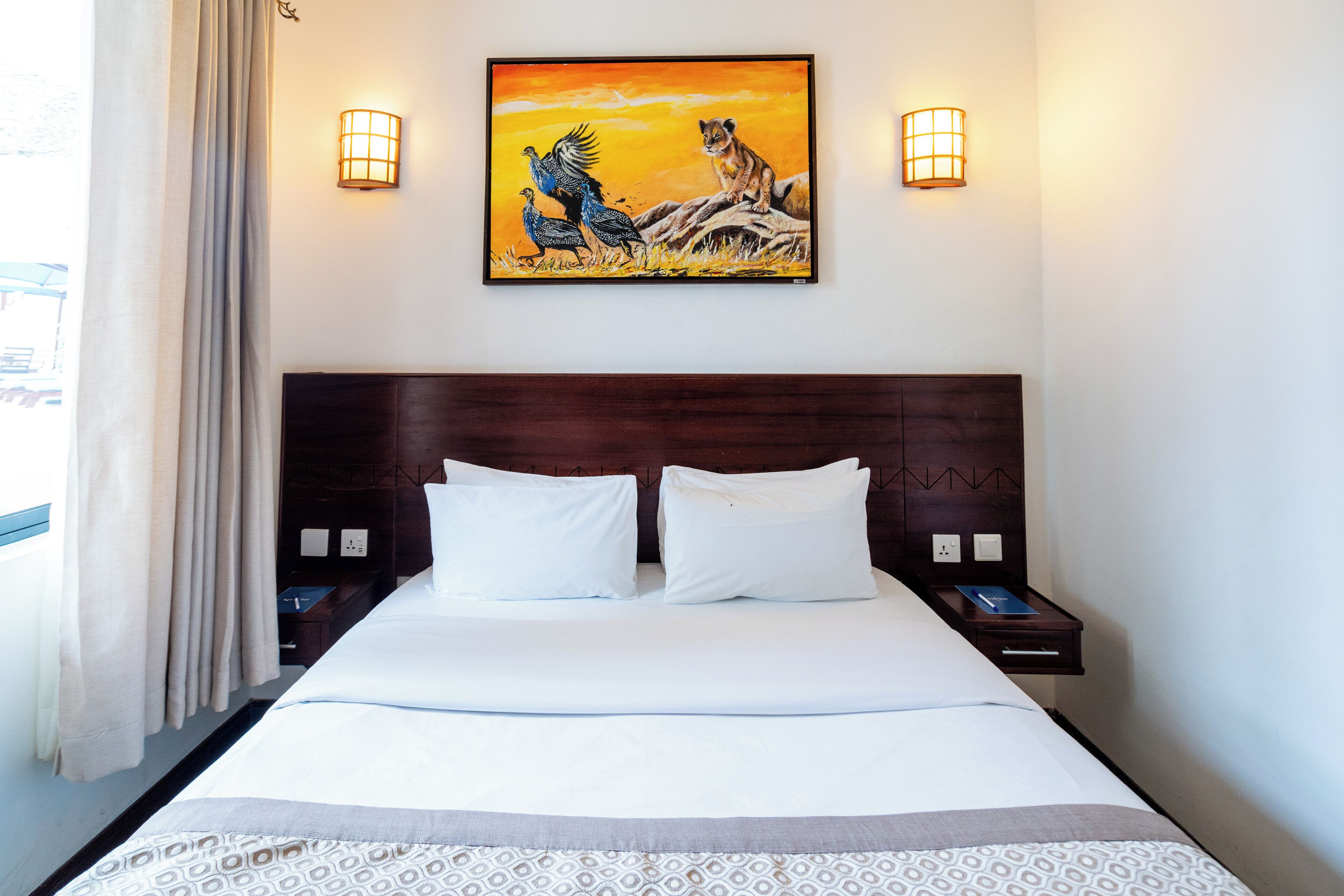 Una habitación de hotel con una cama grande, dos mesitas de noche, una lámpara de noche, un cuadro sobre la cama y una ventana con cortinas.
