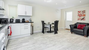 Een moderne keuken met witte kastjes, een eethoek met een tafel en stoelen, en een zithoek met een bank en een rood kussen.