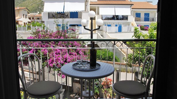 A balcony with two chairs, a table with a bottle and glasses, and a view of buildings with blue shutters. - La Bouganville Studio 200 m From the sea (Sperlonga)