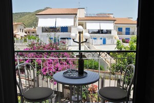 Un balcone con due sedie, un tavolo con una bottiglia e dei bicchieri, e vista su edifici con persiane blu.