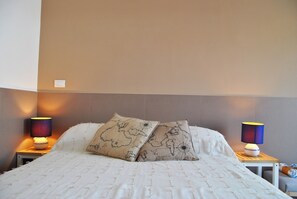 Un lit bien fait, agrémenté de deux coussins décoratifs, de deux lampes de chevet et d’une petite table avec une lampe.