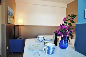 Une chambre d’hôtel avec une table à manger dressée, comprenant des assiettes, une tasse bleue, un vase avec des fleurs violettes, une lampe et une armoire bleue.