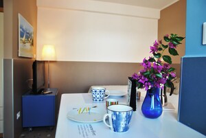 Una camera d'albergo con un tavolo da pranzo apparecchiato, piatti, una tazza blu, un vaso con fiori viola, una lampada e un armadietto blu.