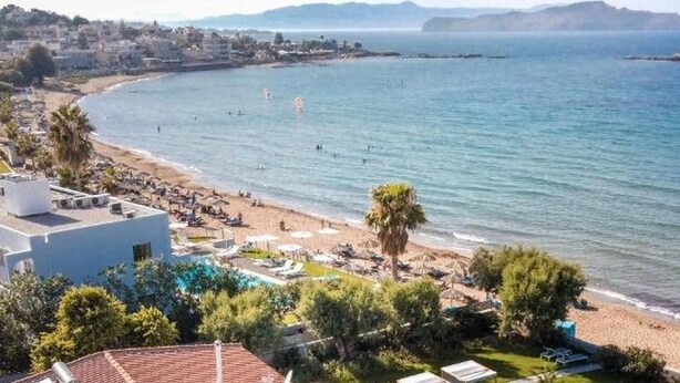 Ett hotell vid kusten med utsikt över stranden, en swimmingpool och en bergskedja i bakgrunden.