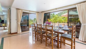 A dining area with a large table set for six, wooden chairs, and a view of lush greenery outside. - Luxury Four Bed Villa Jasmine, Infinity Pool, Full Staff , Chef and Breakfasts (Karon)
