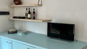 A modern living room with a flat-screen TV, a light blue cabinet, and two wooden shelves holding decorative items and wine bottles. - Villa a few Steps From the Beach (Santa Teresa di Gallura)