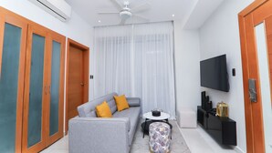 Una sala moderna con un sofá gris, una mesita redonda pequeña, un reposapiés estampado, un televisor montado en la pared y un ventilador de techo.