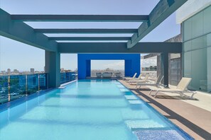 A rooftop infinity pool with a clear view of the city skyline, surrounded by lounge chairs and a blue architectural structure. - Apartamento S1401 - Downtown Santo Domingo (Santo Domingo)