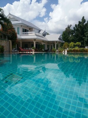 Uma grande piscina com água azul cristalina, rodeada por um edifício com telhado em azulejos e uma área de varanda coberta.