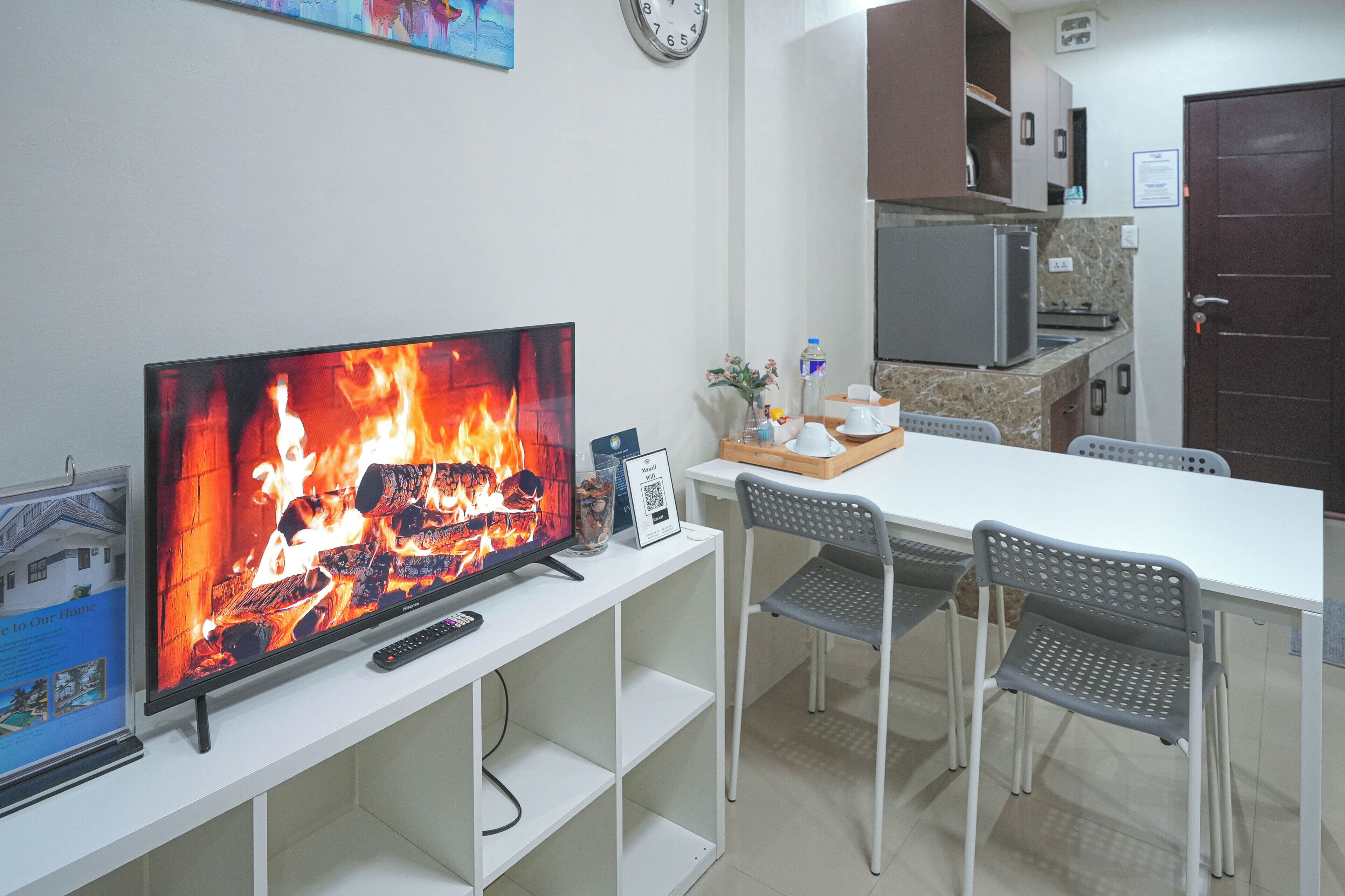 A living room with a flat-screen TV displaying a fireplace, a dining table with chairs, a kitchen area with a refrigerator, and a clock on the wall.