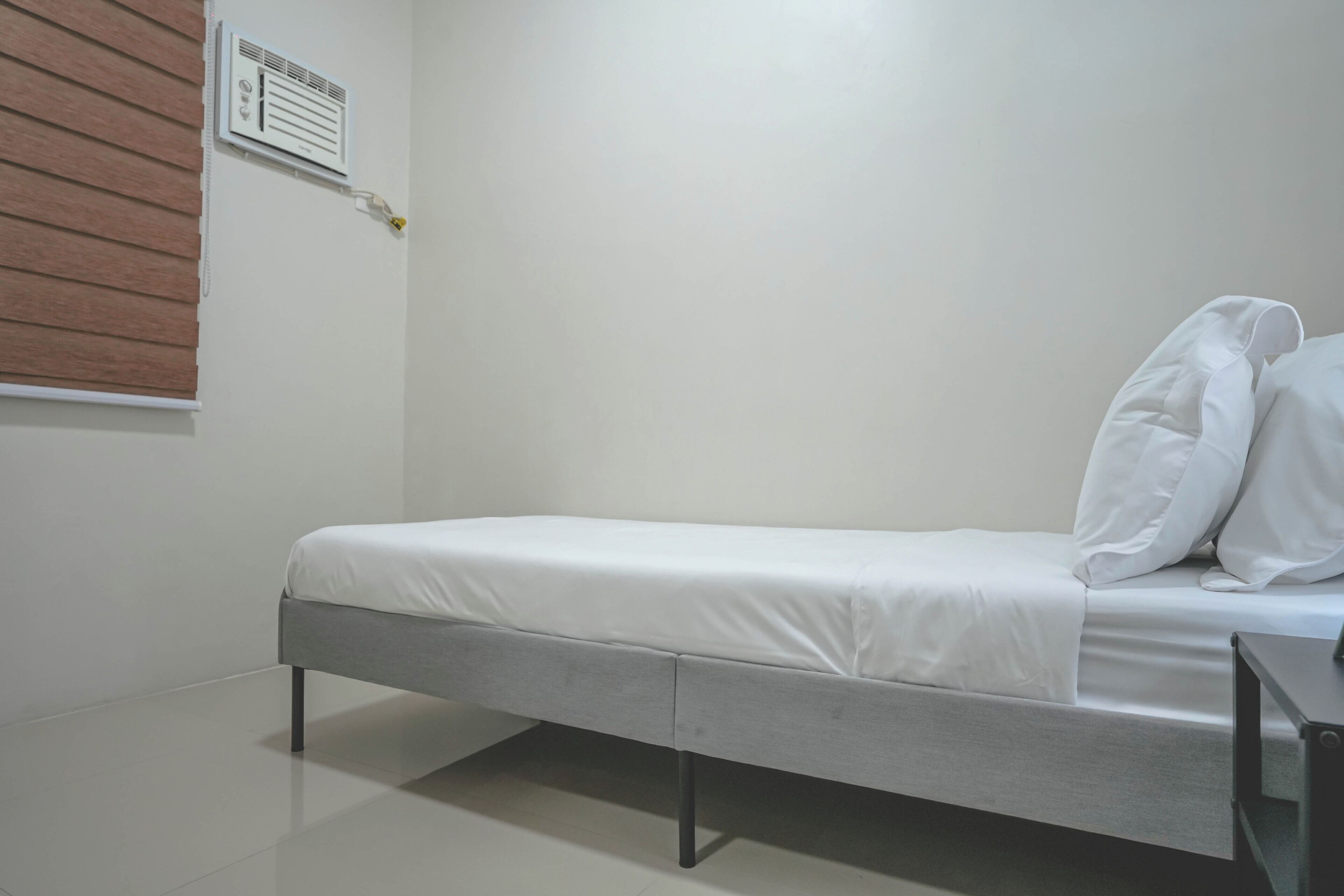 A single bed with white bedding, a wooden slat window blind, and a wall-mounted air conditioning unit.