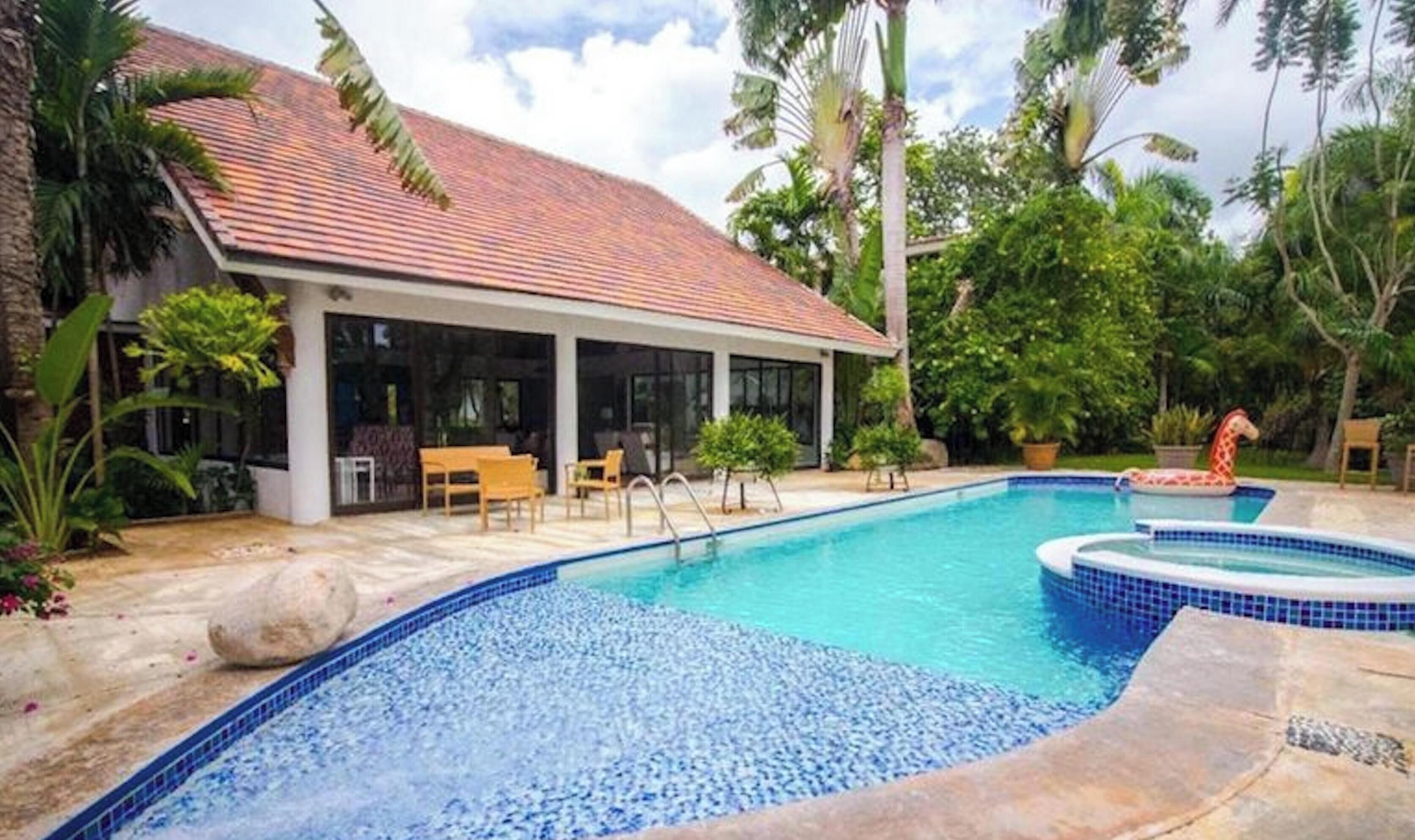 A swimming pool with a tiled deck, a covered seating area, and lush tropical vegetation.