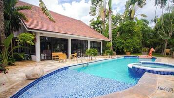 A swimming pool with a tiled deck, a covered seating area, and lush tropical vegetation.