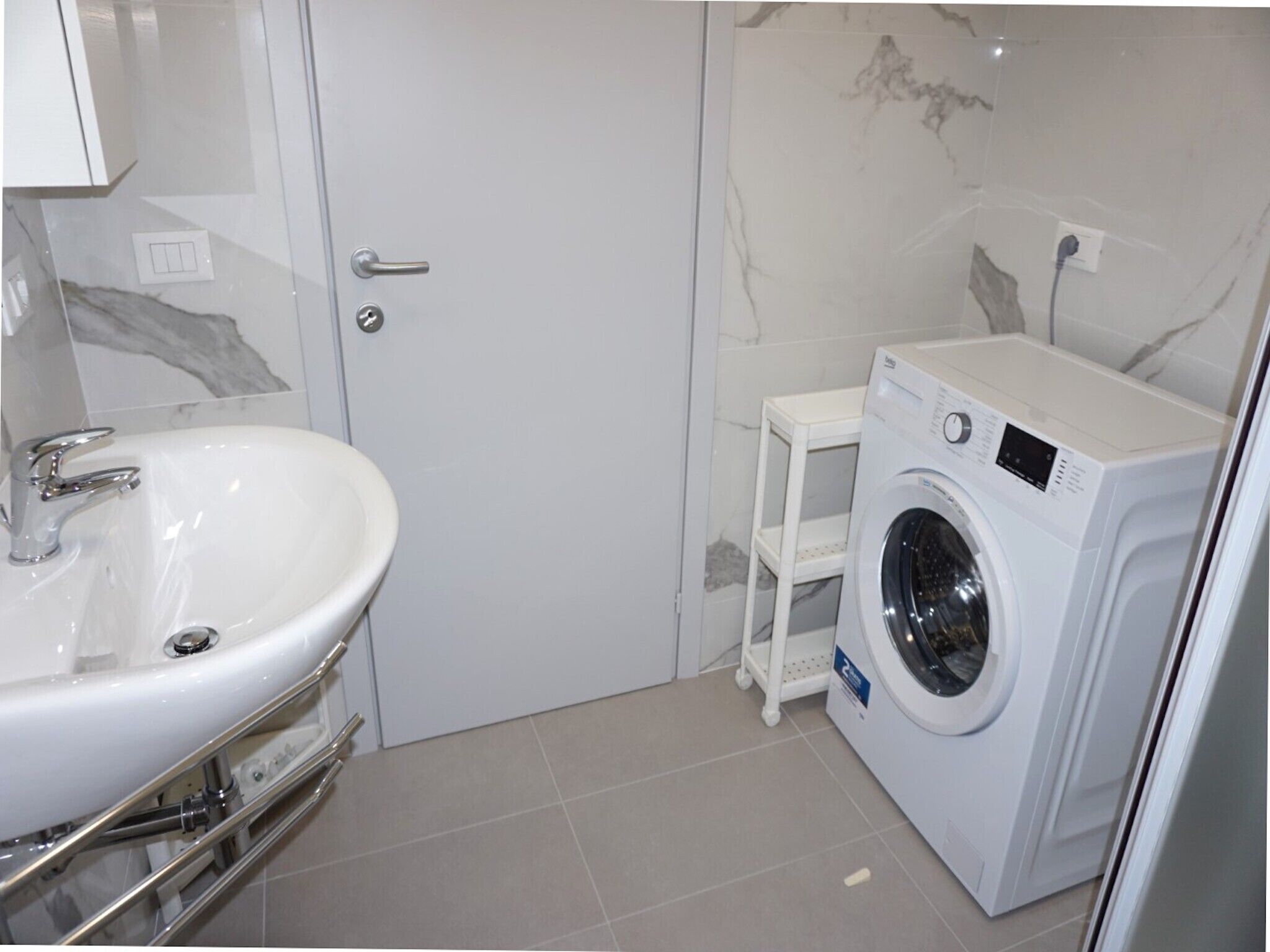 Un baño con un lavabo blanco, una lavadora y un pequeño estante blanco.