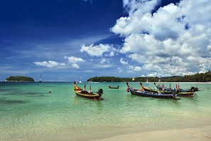 A beach with clear turquoise water, several traditional boats anchored near the shore, and a distant island with a lighthouse. - Kata Beach Ozone Superb Fully Equipped Studio Kitchen Washing Machine Aircon (Karon)