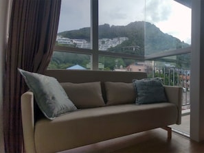 Ein Sofa mit Kissen in einem Raum mit großen Fenstern und Blick auf eine Hügelkuppe mit Gebäuden.