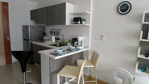 A compact kitchen with a refrigerator, sink, and a small dining area with two chairs. There is a clock on the wall and a fan on the floor. - Kata Beach Ozone Superb Fully Equipped Studio Kitchen Washing Machine Aircon (Karon)