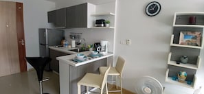 Eine kompakte Küche mit Kühlschrank, Spüle und einer kleinen Essecke mit zwei Stühlen. An der Wand hängt eine Uhr und auf dem Boden steht ein Ventilator.