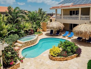 A swimming pool with a hot tub, surrounded by lounge chairs and thatched umbrellas, with a garden and palm trees in the background. - Gorgeous 2-bedroom Apartment 2 km From Eagle Beach (Noord)