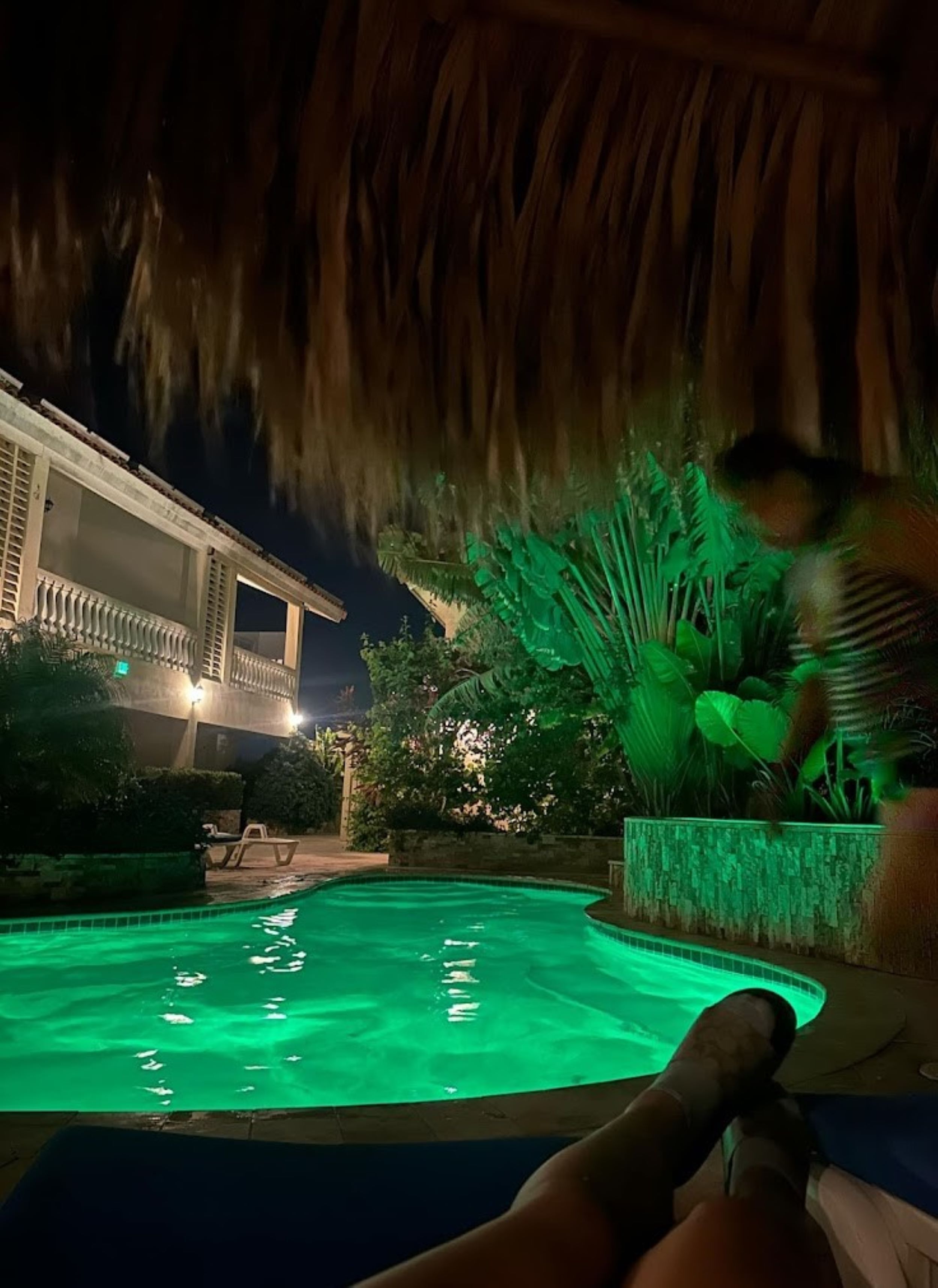 A pool area with a thatched roof structure, a pool illuminated with green lighting, and a building with a balcony in the background.