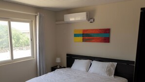 A bedroom with a large bed, a window with curtains, an air conditioning unit, and a colorful abstract painting on the wall.