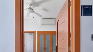 A hallway leading to a room with a ceiling fan, a white sofa with a green pillow, and a wooden door with a number sign. - Apartamento N1401 - Downtown Santo Domingo (Santo Domingo)