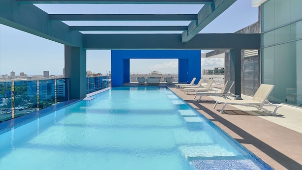 A rooftop infinity pool with a clear view of the city skyline, surrounded by lounge chairs and a blue architectural structure. - Apartamento N1401 - Downtown Santo Domingo (Santo Domingo)