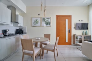 A modern kitchen with white cabinets, a stove, and a dining area with a round table and wooden chairs. There are framed pictures on the wall and a television on the wall.