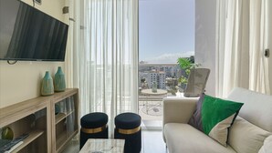 A living room with a flat-screen TV, a sofa with a green pillow, a coffee table, and a balcony with a view of buildings. - Apartamento N1401 - Downtown Santo Domingo (Santo Domingo)